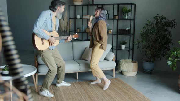 Man Playing the Guitar and Dancing with Woman Who is Singing in Hairbrush Having Fun at Home