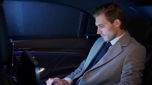 Young Businessman Sitting in a Moving Car and Using Laptop Man Top Manager in a Suit is Sitting in