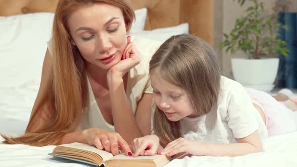 Woman Reading a Book with Her Little Daughter Lying on Bed at Home