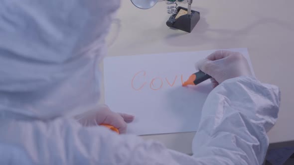 Health Care Worker in Protective Antiviral Suit Writing Covid-19 on a Sheet of Paper