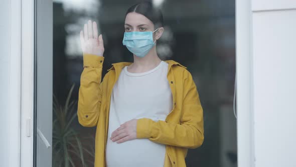 Portrait of Sad Pregnant Woman in Coronavirus Face Mask Looking Out Window Putting Hand on Glass