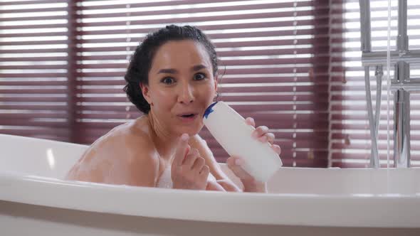 Young Funny Pretty Hispanic Woman Sitting in Bath Bathing Singing Song Using Shampoo Bottle As