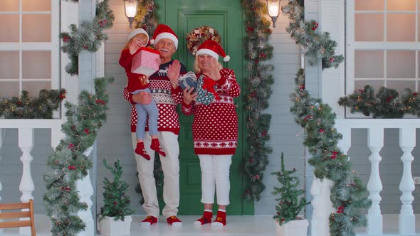 Senior Grandmother Grandfather with Granddaughter Standing at Christmas House Porch Waving Hello Hi