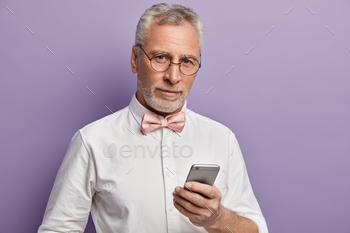 eelancer watches webinar from internet website, holds mobile phone, wears optical glasses, white elegant shirt, models over lilac background.