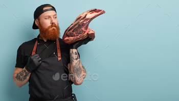 y big red raw piece of meat, needs to chop for preparing dish, wears black cap, t shirt and apron, stands against blue wall with empty space for text