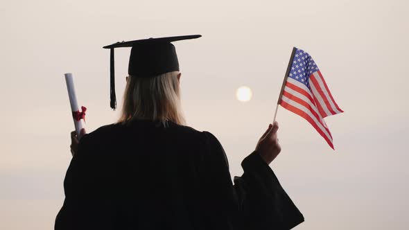 A Student in a Mantle and Cap Holds a Diploma and the Flag of the United States