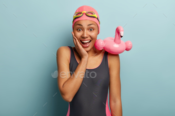 zing, touches cheek, enjoys summer, dressed in swimming costume, has rubber flamingo ring on right shoulder, stands over blue studio background