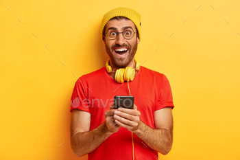 new app for listening music on mobile phone, smiles at camera, types message, dressed in red t shirt, has happy excited look, isolated on yellow