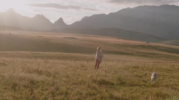 Happy woman with dreadlocks Runs through a Field with playful dog under sunlight in evening.