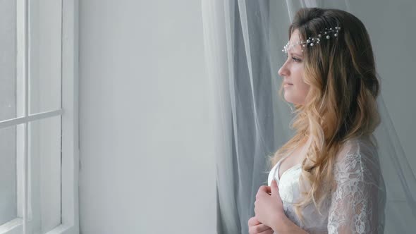 Young Slim Sexy Blond Girl in Lingerie and Boudoir Standing Near Windowsill and Looking in Window.