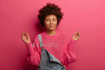  in lotus pose, closes eyes and stands relieved, breathes deeply, controls her feelings, wears knitted sweater and overalls, being stress free