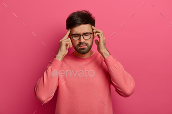 es to relieve pain, suffers from migraine after noisy atmosphere, has thick beard, wears rosy jumper, stands against vibrant crimson background