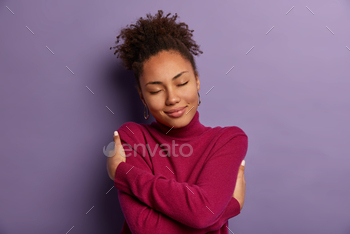 body, hugs herself, closes eyes from pleasure, wears soft turtleneck just for cold weather, feels comfort, stands indoor against purple wall
