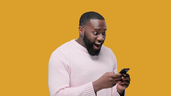 Young Black Man Excited and Celebrating While Holding and Looking at His Smartphone on Yellow