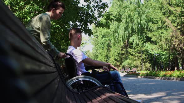 A Woman Brings a Guy in a Wheelchair Next to a Bench and Sits Nearby