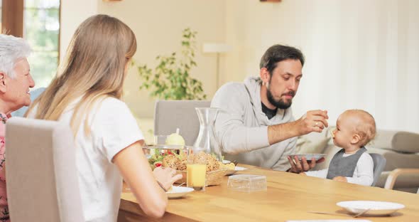 A Brunette Men Sits at the Table with His Parents and Feeds His Little Cute