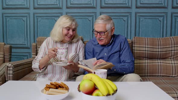 Smiling Senior Couple Grandfather Grandmother Resting on Sofa Drinking Coffee Reading Book at Home