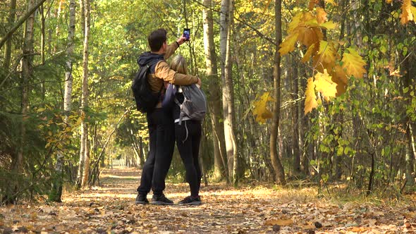 A Hiking Couple Walks Through a Forest on a Sunny Day, Stops To Take a Selfie with a Smartphone