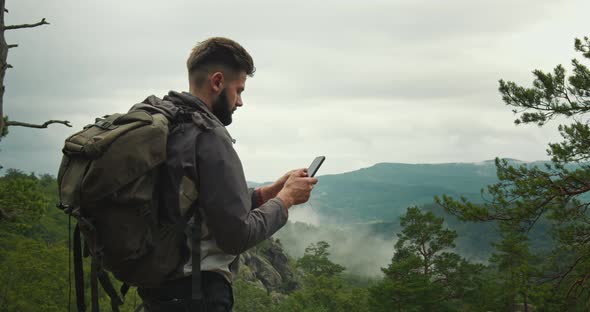 A Man is Standing on Top of a Mountain and Typing a Message on His Smartphone