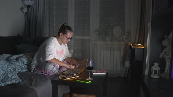 Woman in Pajamas and Glasses is Sitting on Sofa and Working on Laptop at Home