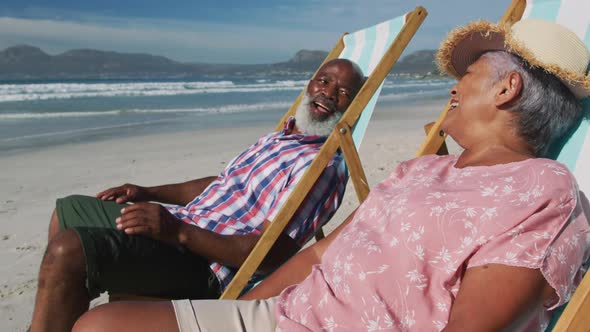 Senior african american couple sitting on sunbeds and smiling at the beach