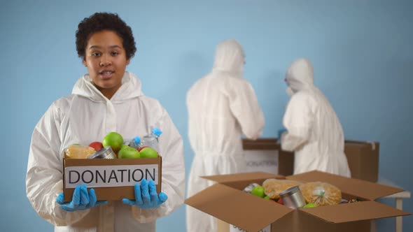 Young African Woman in Protective Overall and Gloves Holding Box with Food for Charity