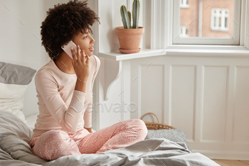 tisfied with tariffs and internet connection at home, wears casual nightwear, looks aside, at window, sits on bed, discusses something with friend