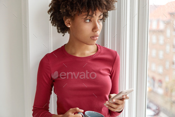 ands near windowsill, drinks aromatic beverage, holds modern cell phone, waits for important call, wears casual red sweater, looks out of window