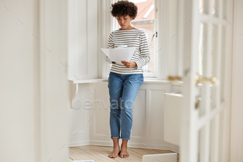 an studies provision for taxation, holds papers, dressed in casual clothes, stands near window sill in spacious light room, ratifies contract