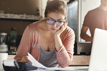 ssed wife holding piece of paper and calculating finances, trying her best to cut domestic expenses using calculator and generic laptop computer