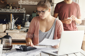 alculating expenses while doing family budget using generic laptop and calculator at home, her husband standing on background with mobile phone