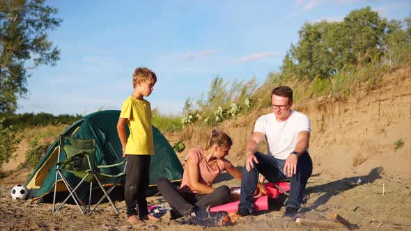 Young Family with a Child is Warming Around a Fire and Tents on Nature in Summer