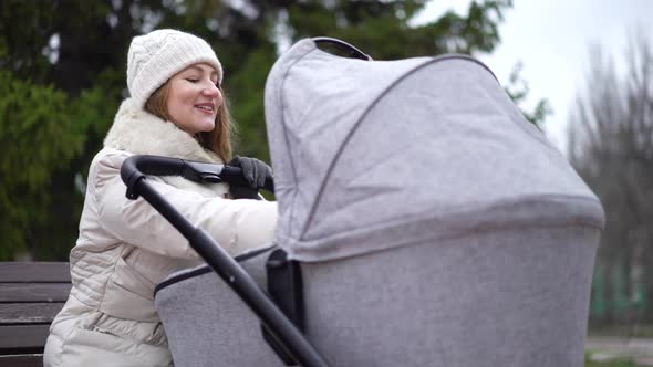 Young Mother with Newborn Child Outdoor. She Sitting on the Bench with Baby Sleeping in Pram