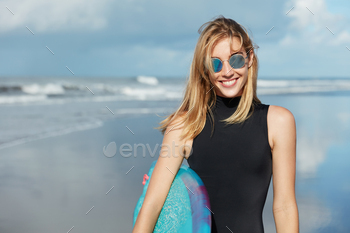 t. Smiling beautiful young woman in shades and swimsuit, holds surfboard, poses against ocean background, being in good mood as likes her hobby