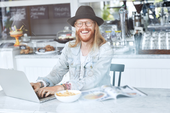 dish beard and mustache, wears glasses, trendy hat, enjoys lunch break, gets ready to work hard, keyboards on laptop computer, connected to wifi