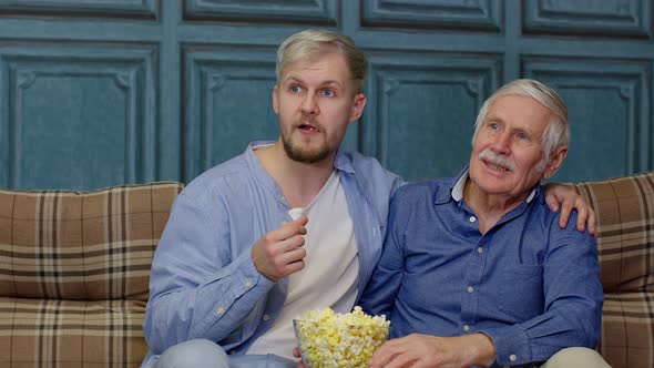 Family Senior Man Father and Adult Son Sitting on Sofa Watching Sport Game on TV with Eating Popcorn