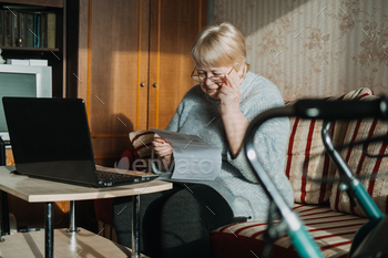 y for Seniors. Mature woman in glasses with Laptop and documents on the background of Rollator Mobility Walker for Seniors at home