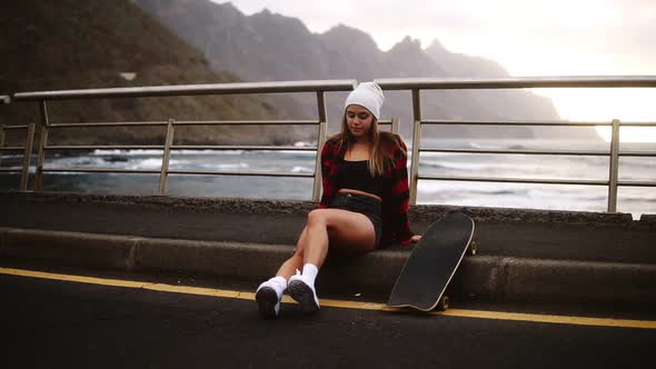 Girl Sitting with Longboard at the Border at Coastline Asphalt Road on Background Hills and Foggy