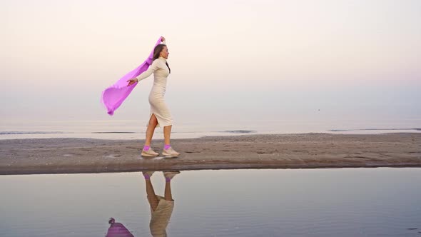 Woman in White Midi Dress Walks on Beach Waving Purple Scarf