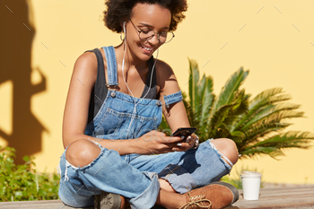 Carefree African American woman searches radio station, enjoys good sound, types email on mobile pho