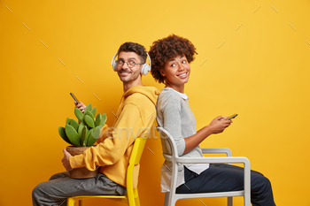 n technologies sit back to each other on chairs hold mobile phones dressed casually enjoy online communication isolated over yellow studio wall