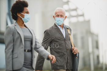 k female colleague using protective mask and talking while walking in front of the office building during COVID-19 pandemic.