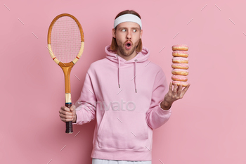  has break during tennis game holds racket and pile of doughnuts leads active life wears headband and sweatshirt isolated on pink background