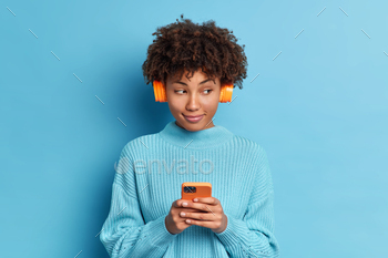 expression listens favorite track in headphones holds modern cellular and sends text messages wears casual jumper isolated over blue studio wall