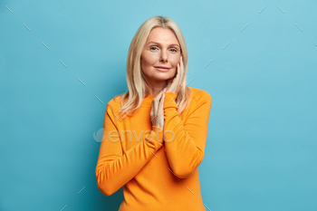ace and enjoys soft skin feels relieved calm looks seriously at camera dressed in casual orange jumper takes care of complexion isolated on blue wall