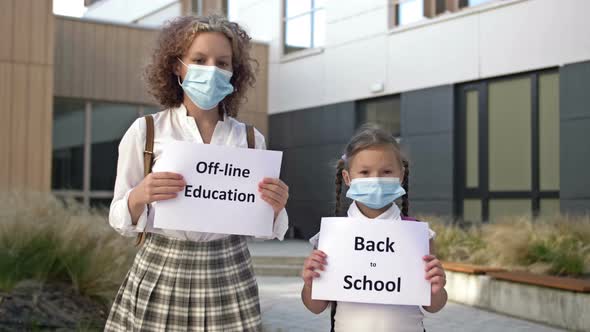 Two Schoolgirls Wearing Medical Masks with Placards Stand in Front of the School Building