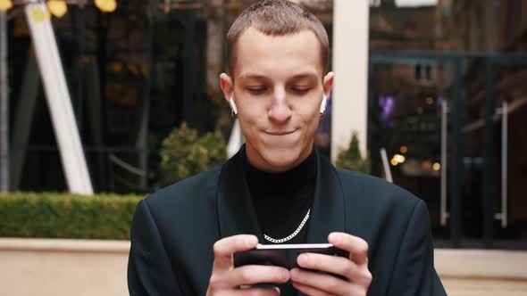Front View of a Happy Man Walking Towards Camera Using a Smartphone, Playing or Chatting. Outside