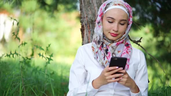 A Muslim in Hijab Communicates on the Phone with Friends, She Sits in a Park