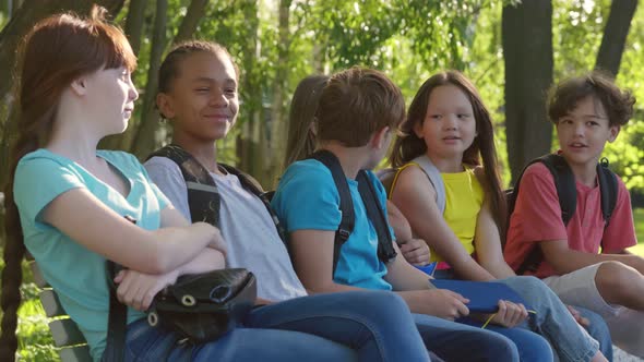 Group of Happy Diverse School Students with Backpacks Sitting on Bench and Talking Outdoors