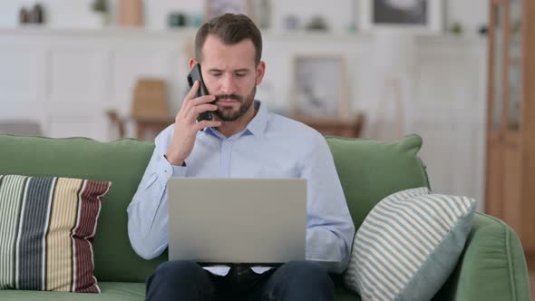 Young Man with Laptop Talking on Smartphone on Sofa 